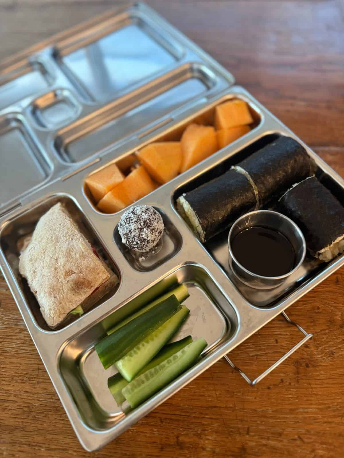 Packed stainless steel lunchbox with sandwich, sushi rolls, cucumber, bliss ball and rockmelon